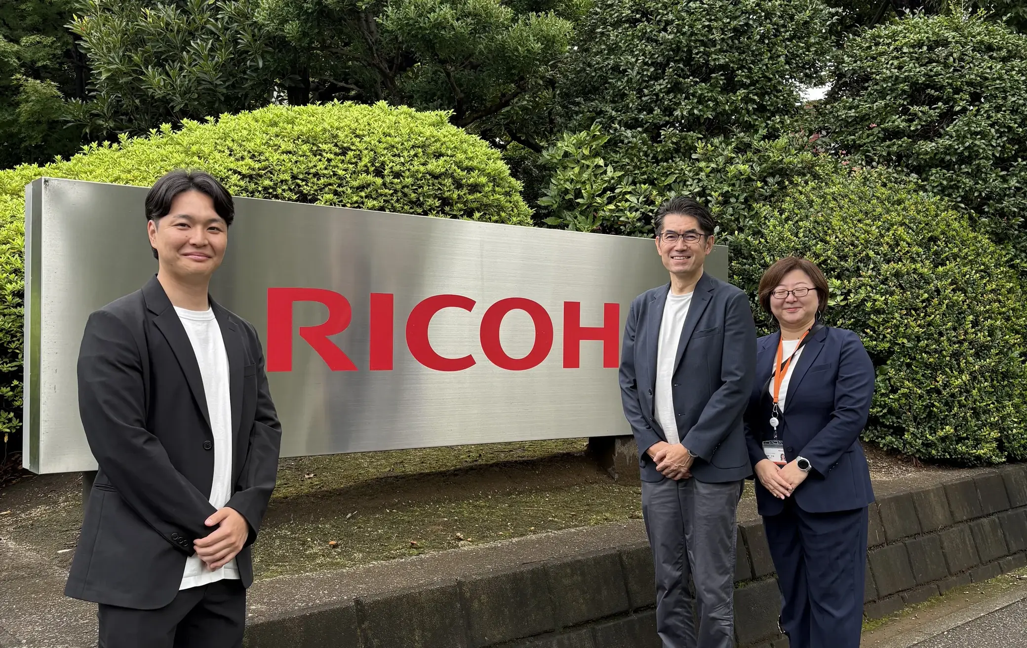 RICOHの社名看板の前に並んで立つ3名